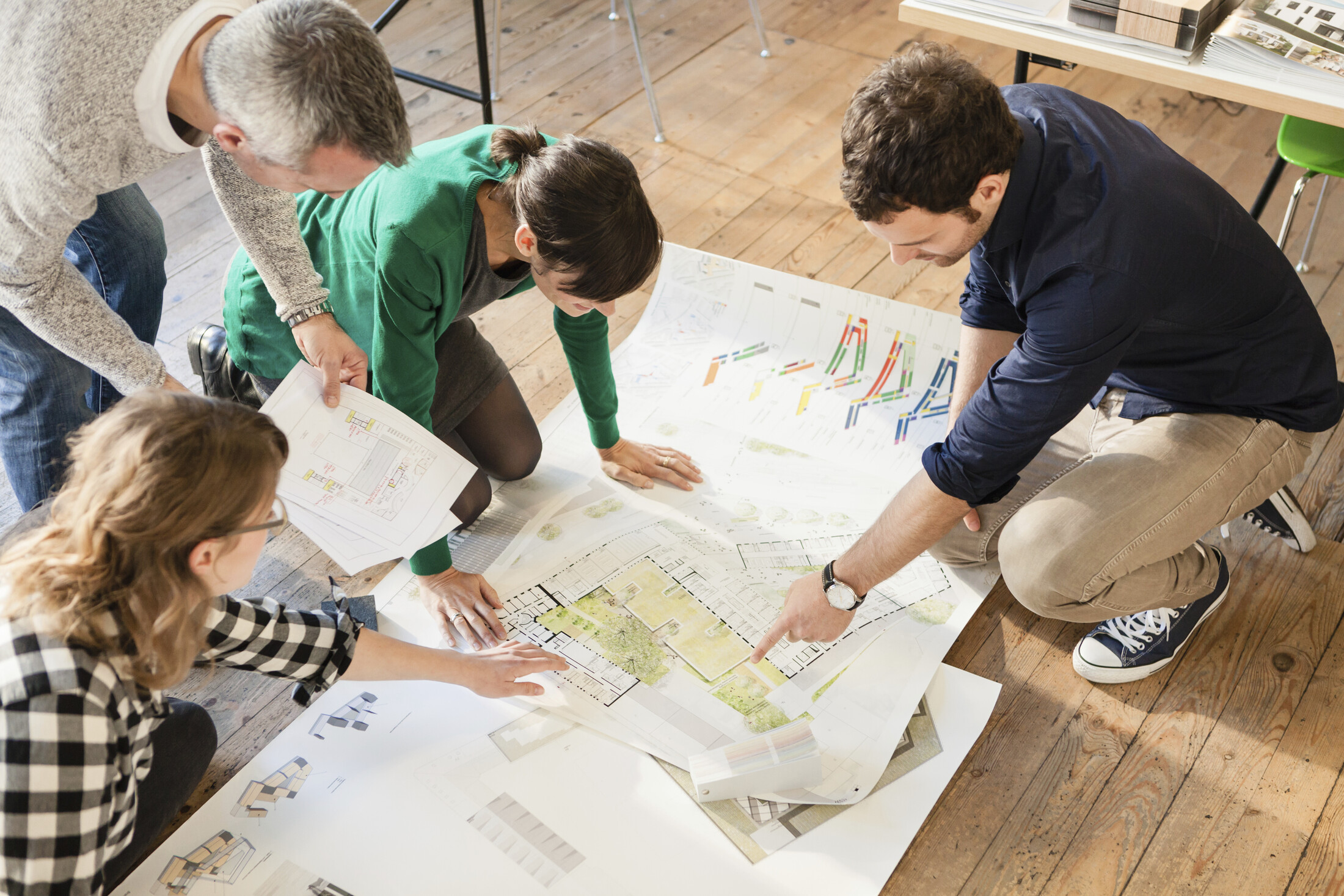 Vier Personen begutachten Großküchen-Baupläne am Boden in einem hellen Raum mit Holzfußboden.