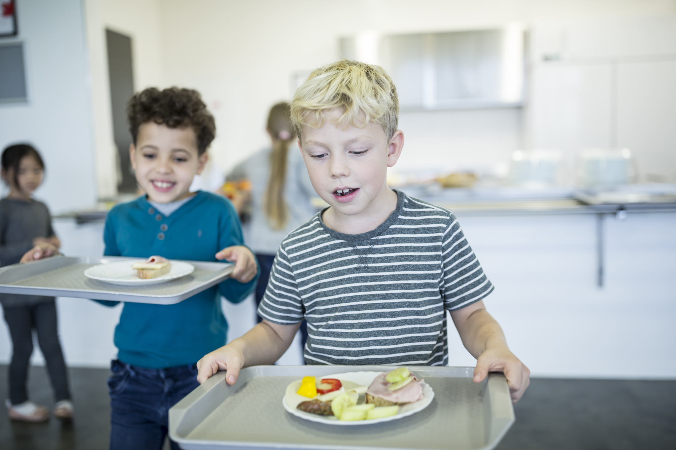 Zwei Jungen tragen Tabletten durch eine Großküchen-Cafeteria, im Hintergrund sitzen weitere Kinder an Tischen.
