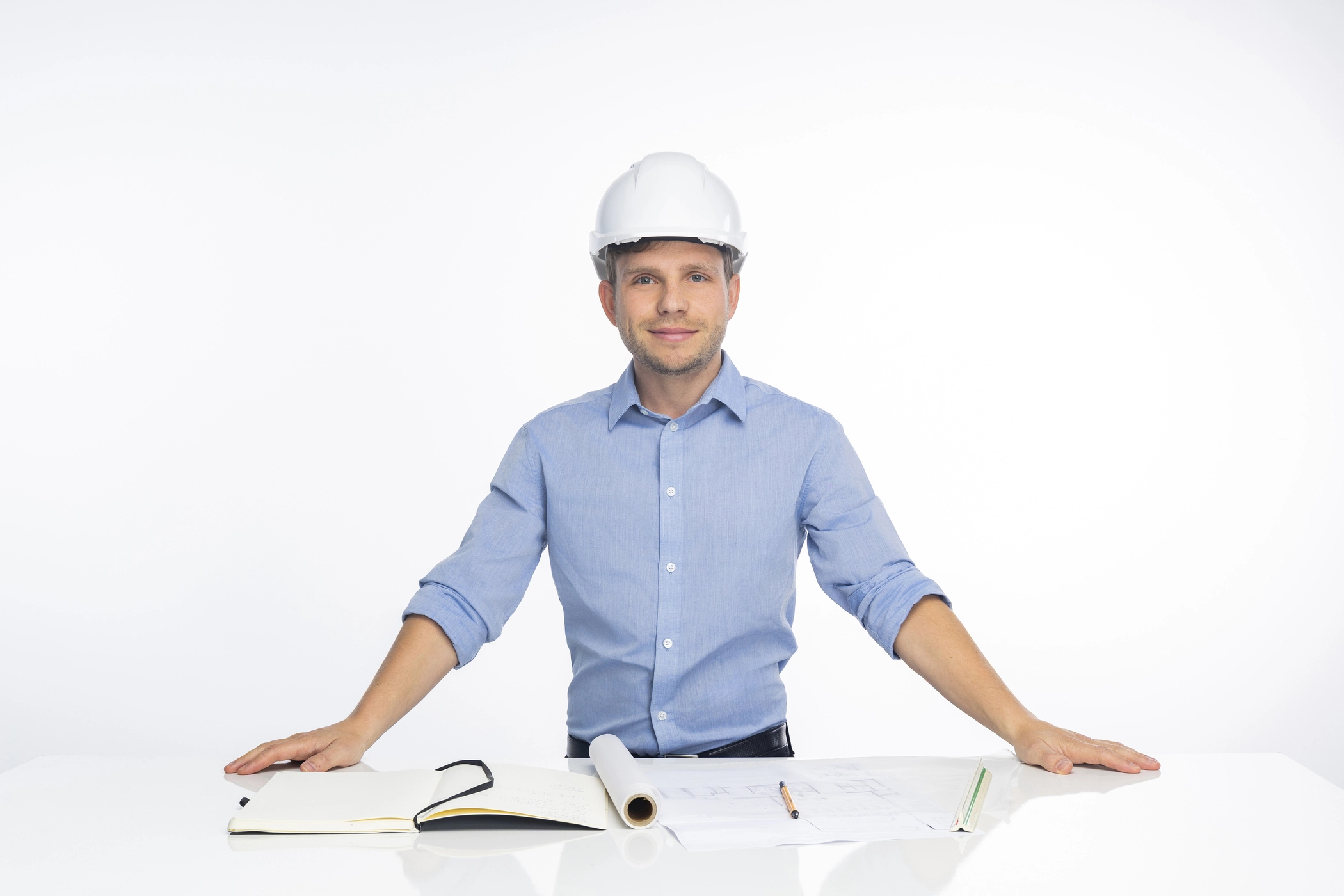 Mann mit Schutzhelm sitzt am Schreibtisch, prüft Großküchenpläne und notiert etwas im Notizbuch.