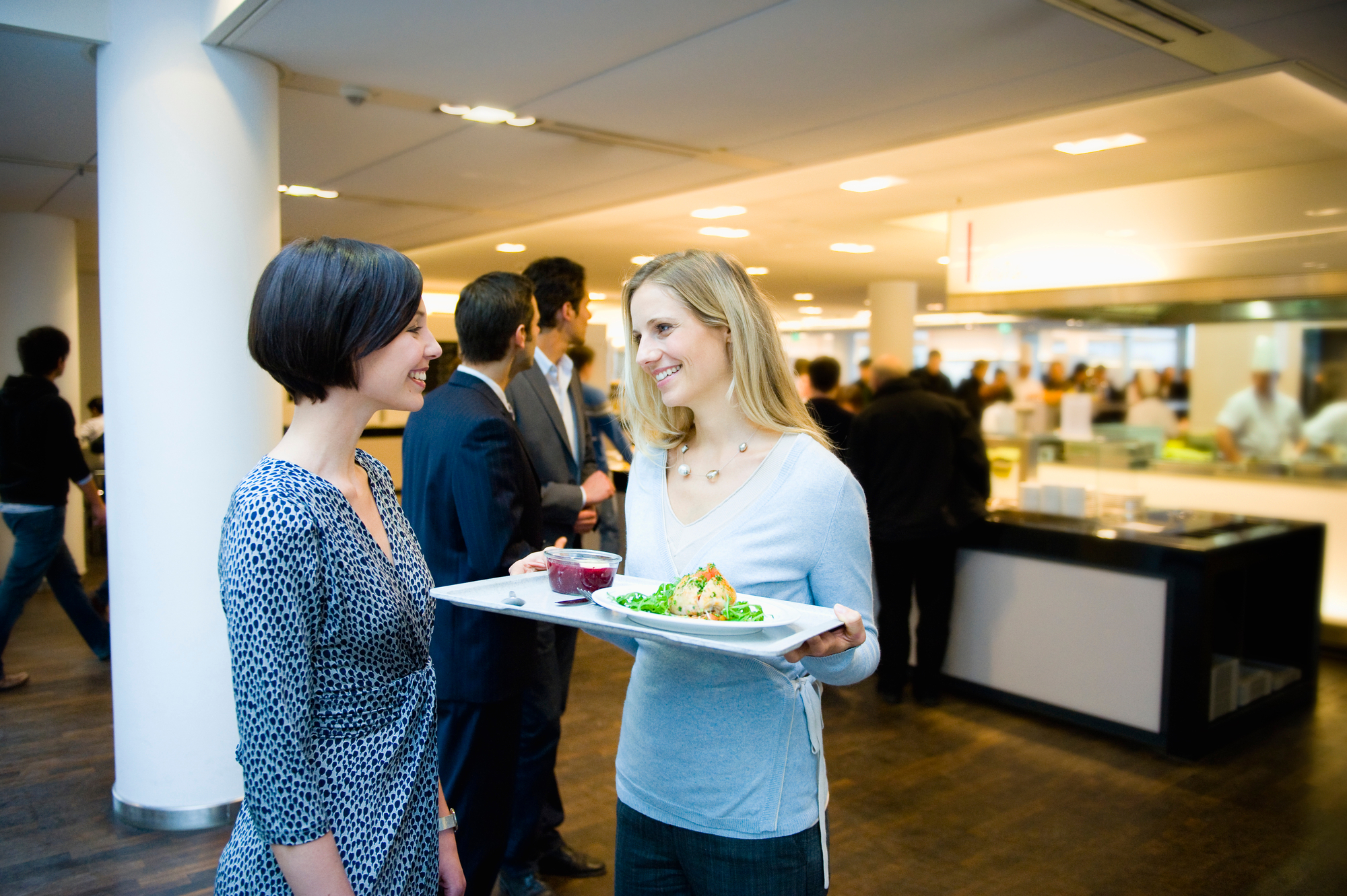 Zwei Frauen unterhalten sich in einer Großküchen-Cafeteria, eine hält ein Tablett mit Essen, im Hintergrund Menschen und Stationen.
